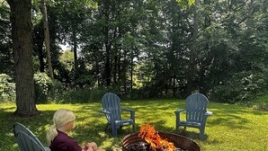 A person seated on a chair by a campfire in a grassy outdoor area with trees in the background. - Bear Lake | Hot Tub, Kayaks, Firepit, Lake & Beach (Lake)