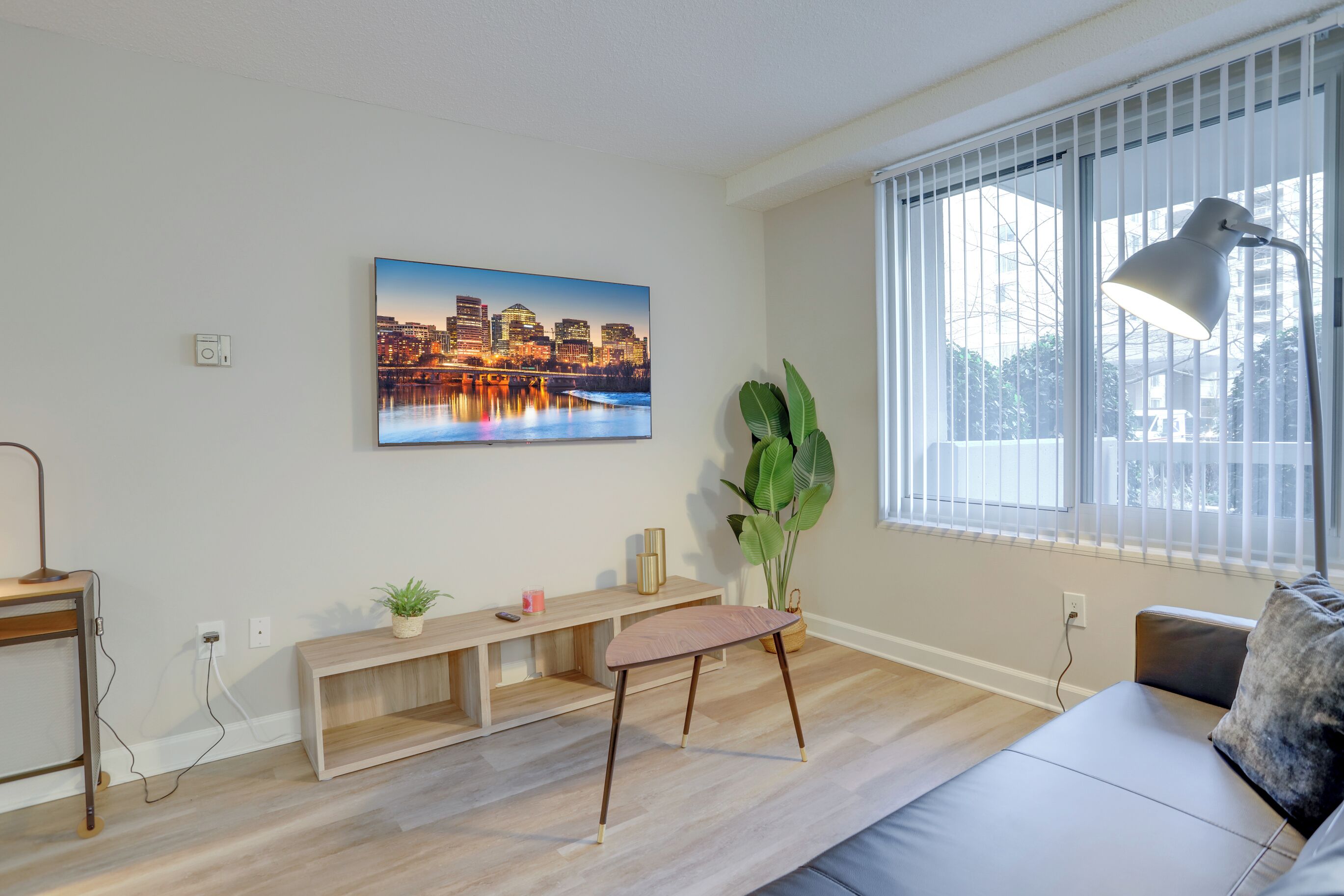A modern living room with a cityscape painting, a large window with blinds, a floor lamp, a sofa, and a wooden console table with a plant.