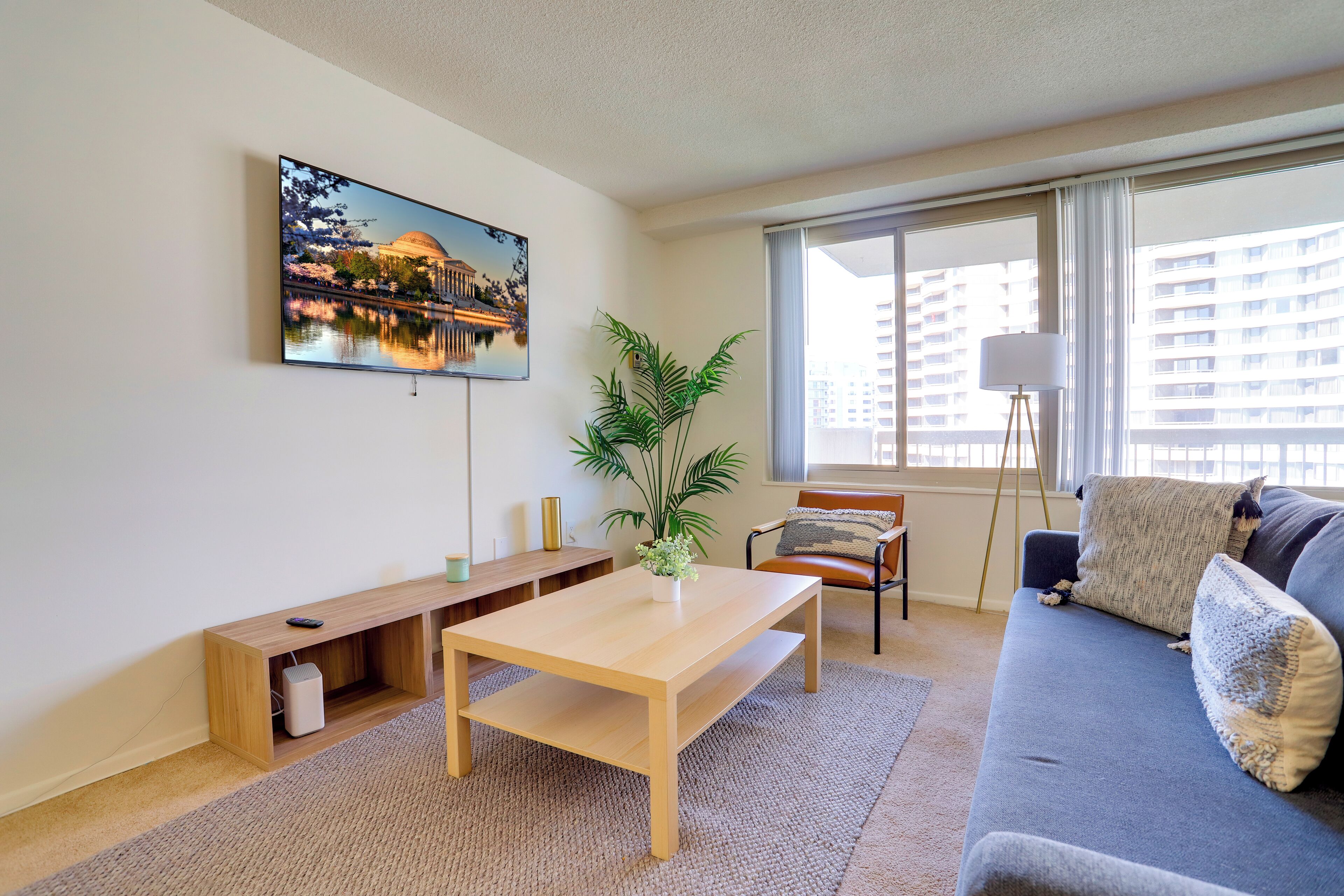 A living room with a sofa, a coffee table, a TV mounted on the wall, a plant, and a window with a view of buildings.
