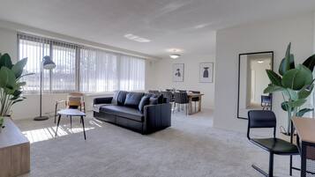 Una sala de estar moderna con un sofá azul oscuro, una mesa de comedor con sillas y grandes ventanales con persianas.