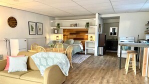 Un salon confortable avec un canapé, une table à manger et des chaises. On aperçoit, au fond, un coin cuisine avec une cuisinière et un évier.