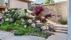 Une maison moderne dotée d’un mur de soutènement en pierre, d’une terrasse en bois et d’un petit jardin agrémenté de diverses plantes et d’un arbre.