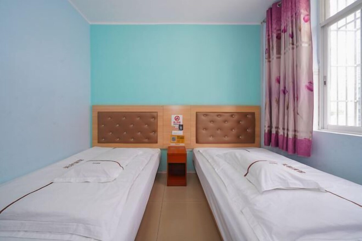 Een kamer met twee eenpersoonsbedden, een houten hoofdbord, een klein houten nachtkastje en een raam met roze gordijnen.
