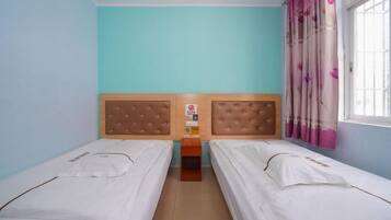 Ein Zimmer mit zwei Einzelbetten, einem Holzkopfstück, einem kleinen Nachttisch und einem Fenster mit rosa Vorhängen.