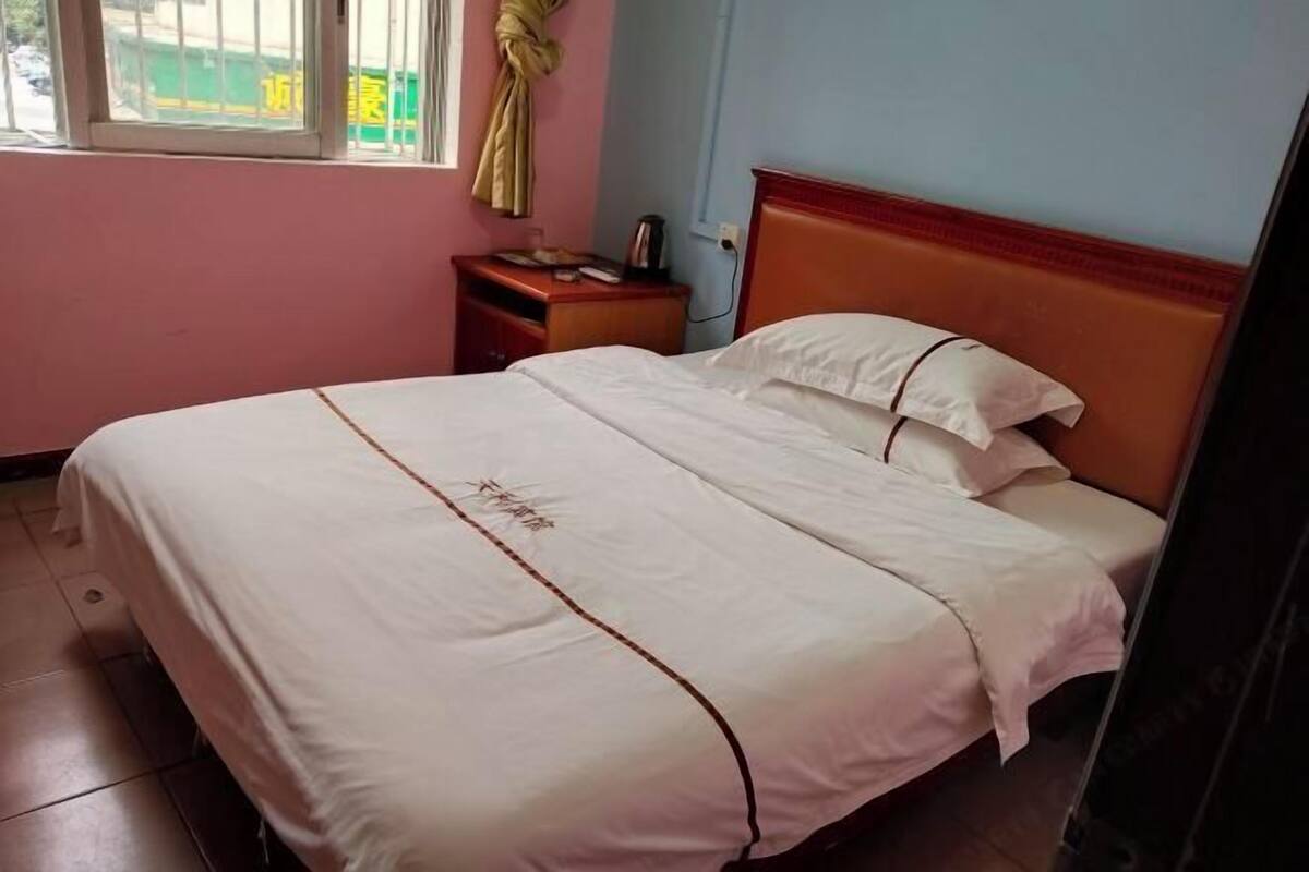 Een net opgemaakt bed met een witte spreideken en kussens, een houten hoofdbord, een klein nachtkastje met een lamp en een raam met gordijn.