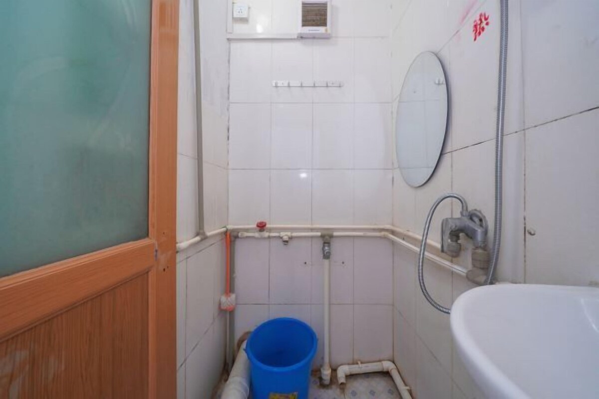 Een kleine badkamer met een wit wastafel, een spiegel en een douche. Er staat een blauwe emmer op de vloer.