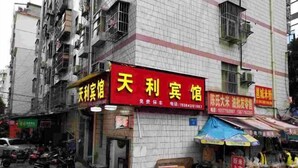 Ein Gebäude mit mehreren Schildern, darunter eines für das Unternehmen „天利宾馆“ und ein weiteres für „陈家大木“. Auf der Straße sind geparkte Fahrzeuge und einige Personen zu sehen.