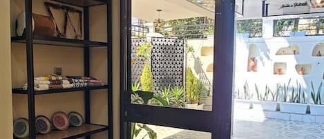 Una habitación con piso de baldosas, una silla de mimbre, un estante de marco negro con objetos decorativos y una puerta que da a un espacio exterior con plantas en macetas y piso estampado.