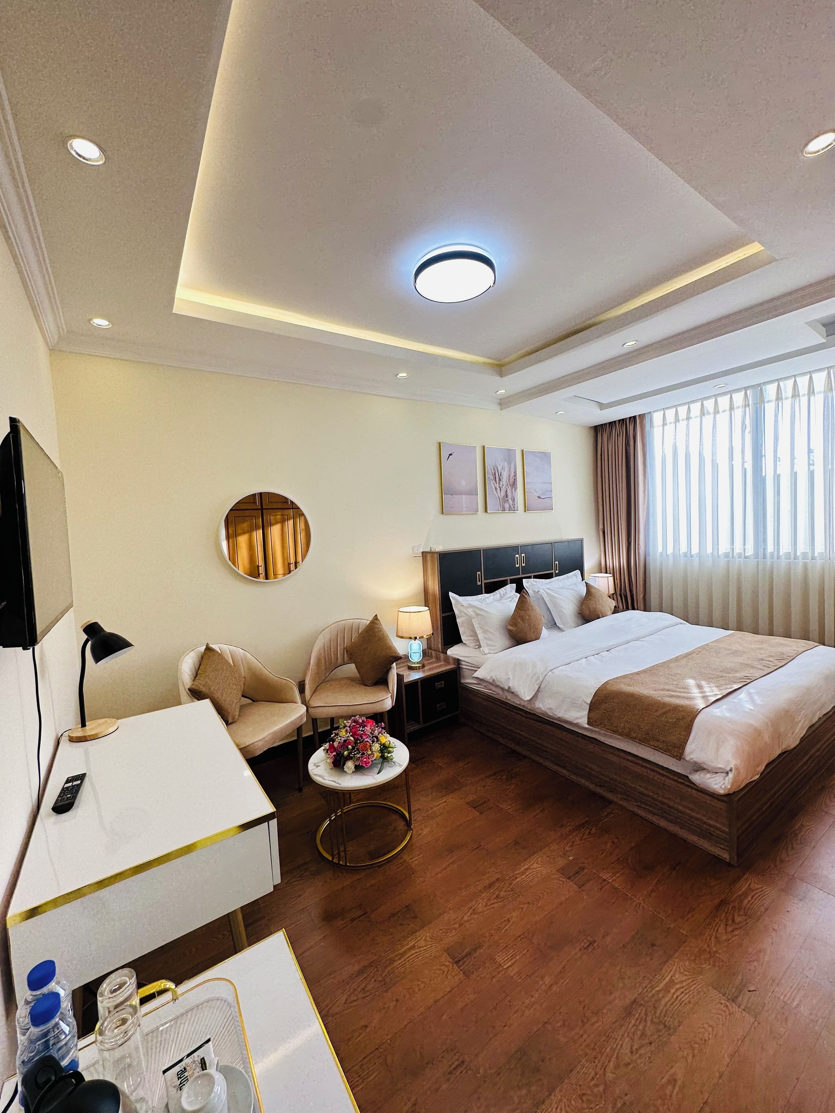 A hotel room with a bed, a desk, a chair, a small table with a vase of flowers, a bedside table with a lamp, and a large window with curtains.