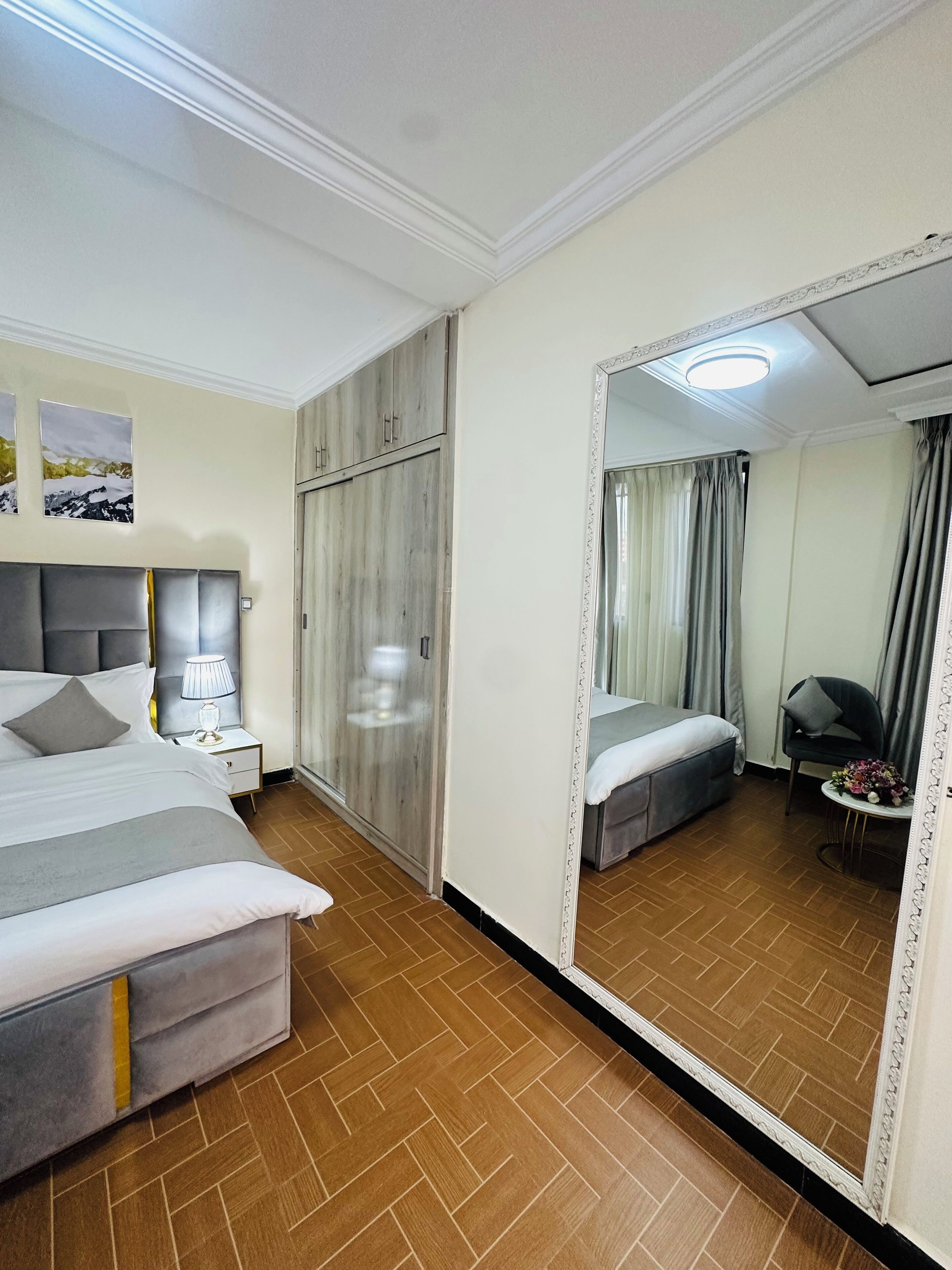 A hotel room with a large bed, a wardrobe, a mirror, and a small table with a flower arrangement.