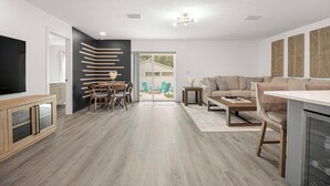 A modern living room with a sofa, coffee table, and TV. There is a dining area with chairs and a table. The room has wooden flooring and a large window. - Santos Place -New Built Home -4Mins To Trailhead- Centrally Located! (Ocala)