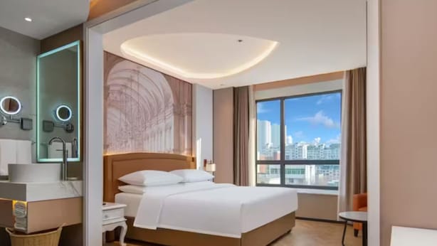 A modern hotel room with a large bed, a vanity with a mirror, and a view of the city through a large window. - Vienna Hotel (Jinan Huayuan Road ) (Jinan)