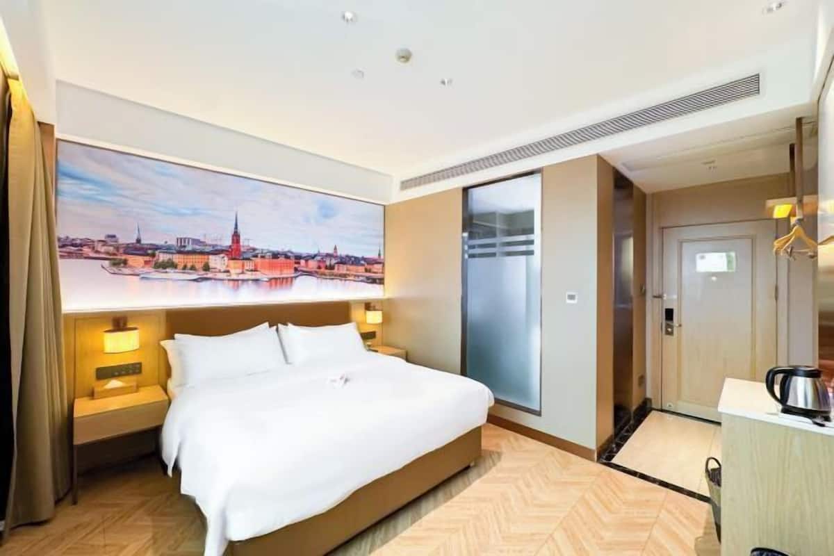 Une chambre d’hôtel comprenant un lit, une table de chevet, une lampe fixée au mur, une grande fenêtre et une porte donnant sur une salle de bain.