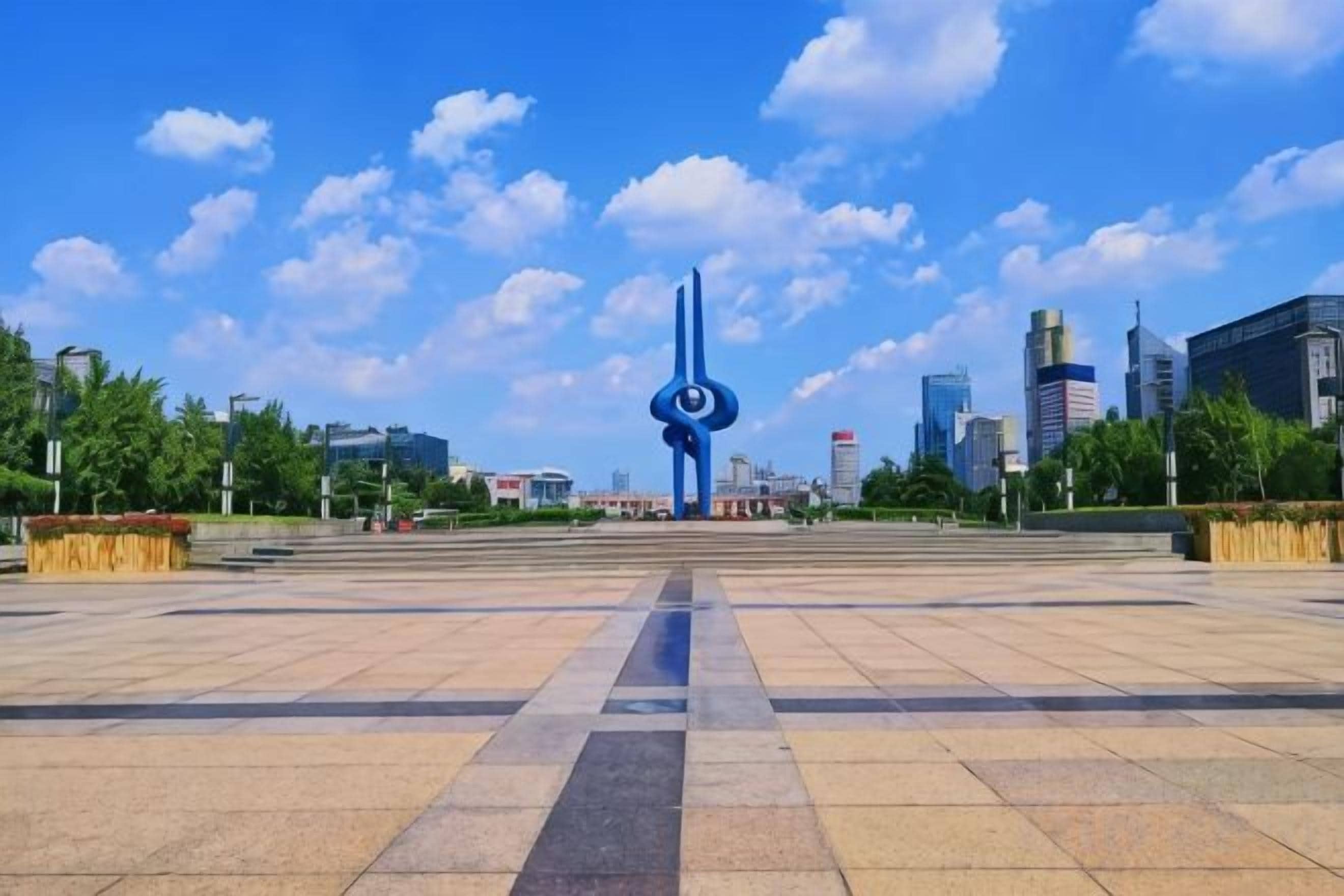 Una amplia plaza con una escultura central, rodeada de edificios modernos y cielos despejados.