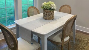 Un coin repas avec une table blanche, quatre chaises en bois, un lustre orné de lampes orange, un panier en osier rempli de fleurs et un miroir en forme de soleil accroché au mur.
