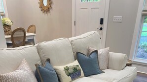 Un canapé d’angle blanc agrémenté de plusieurs coussins, un tapis tissé et un coin repas avec des chaises en osier et un panier de fleurs.