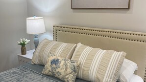 Un lit bien fait, avec une couverture grise, une tête de lit beige et plusieurs oreillers. Une lampe et un petit vase avec des fleurs sont posés sur la table de chevet, à côté du lit.