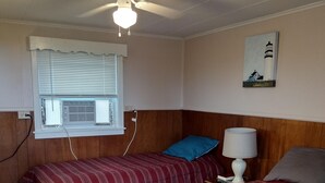 Una habitación con dos camas, un ventilador de techo, una ventana con persianas, una lámpara de pared y un cuadro de un faro.