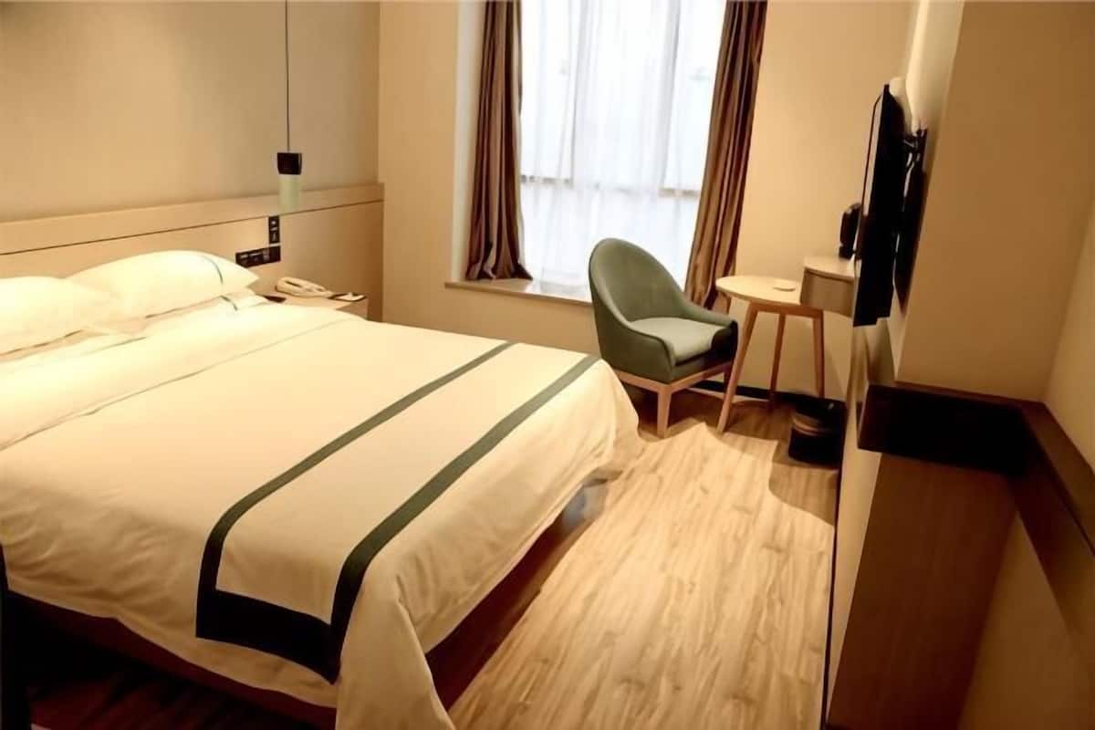 A hotel room with a bed, a chair, a small table, a TV, and a window with curtains.