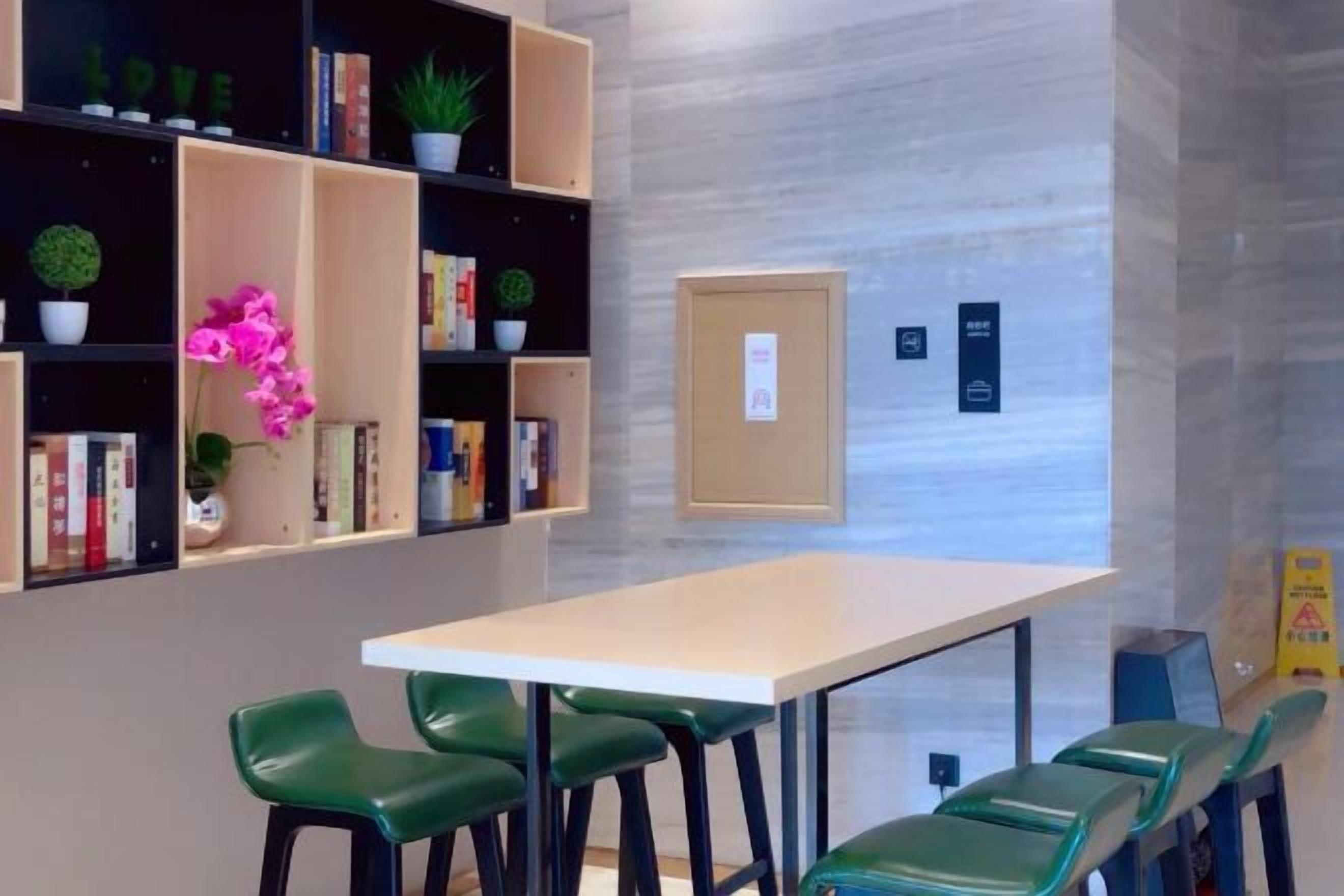 A room with a long table and green chairs, a bookshelf with books and plants, and a bulletin board.