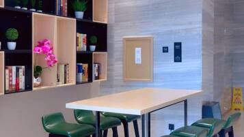 A room with a long table and green chairs, a bookshelf with books and plants, and a bulletin board.