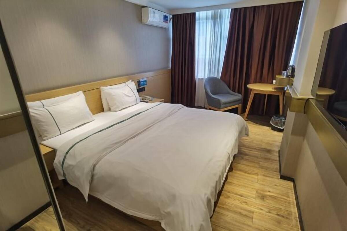 A hotel room with a bed, a chair, a small table, a TV, and an air conditioning unit.