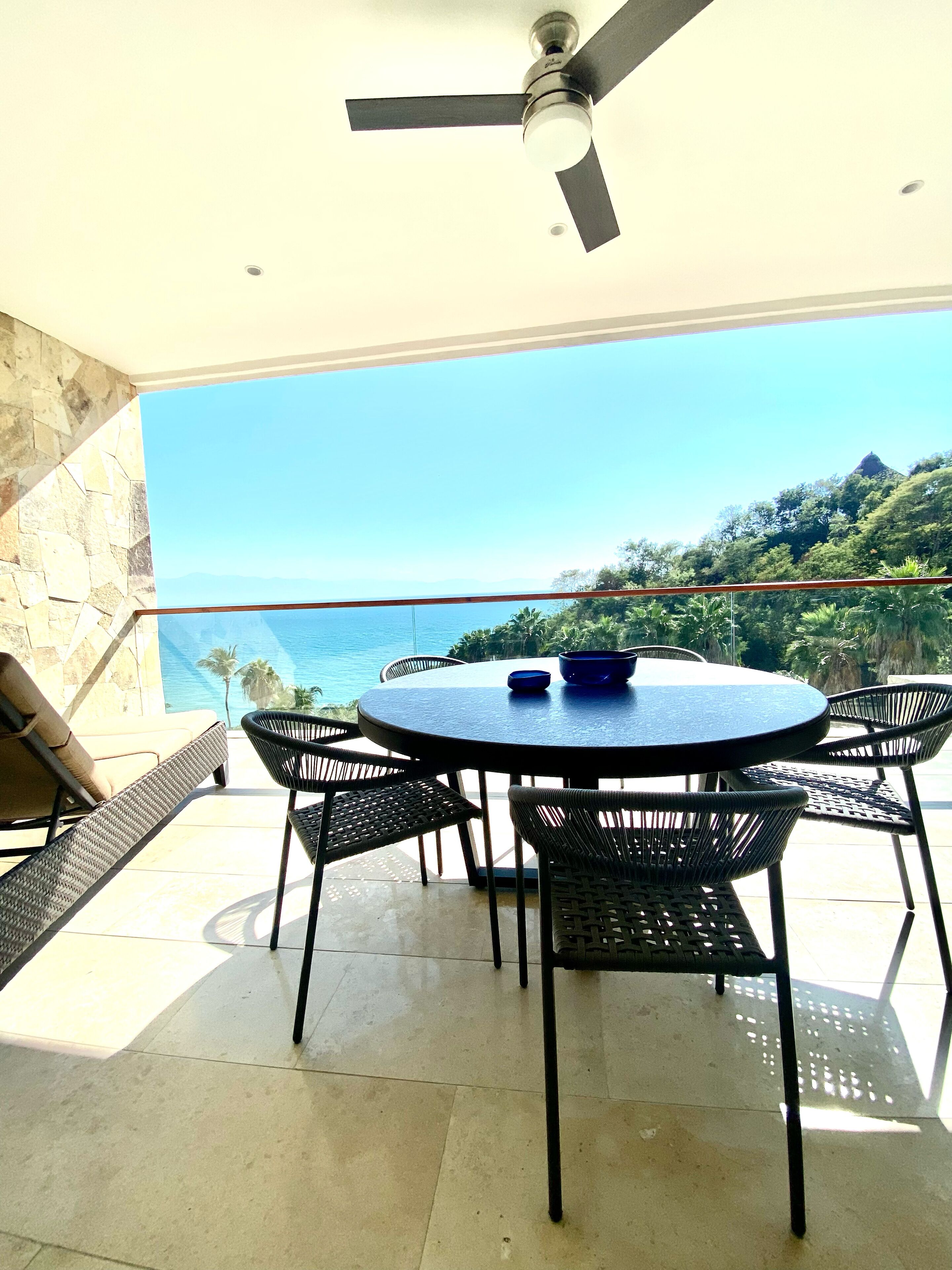 A round table with chairs on a balcony overlooking a scenic view of the ocean and hills.