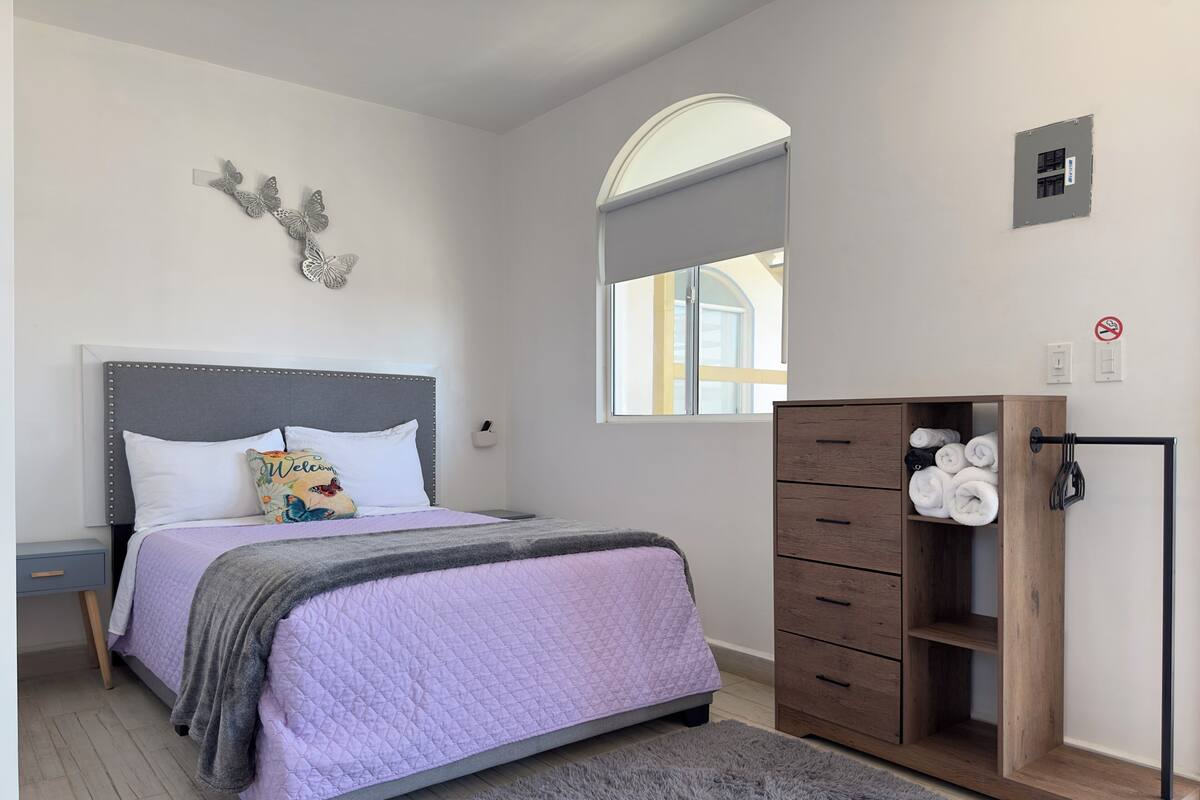 A bedroom with a bed, a nightstand, a wardrobe, a fluffy rug, and a window with a view of a building.