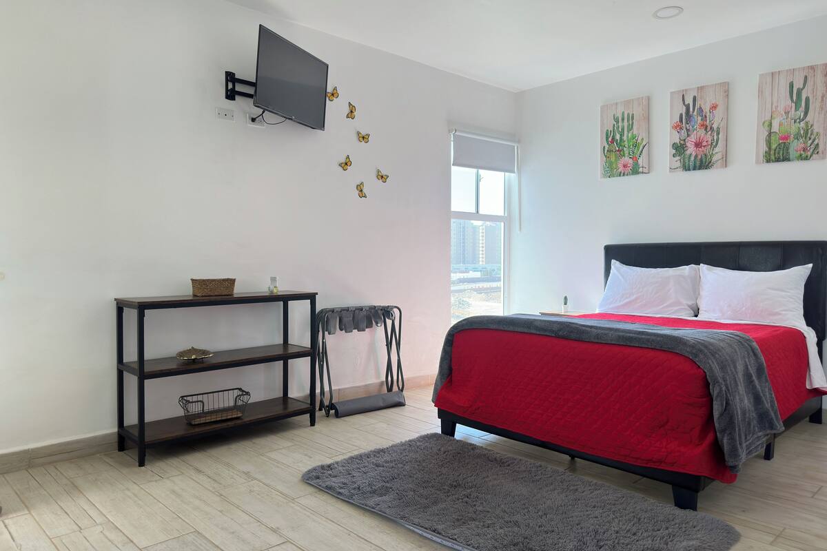 A bedroom with a bed, a TV mounted on the wall, a small table with a basket, and a window with a view of buildings.