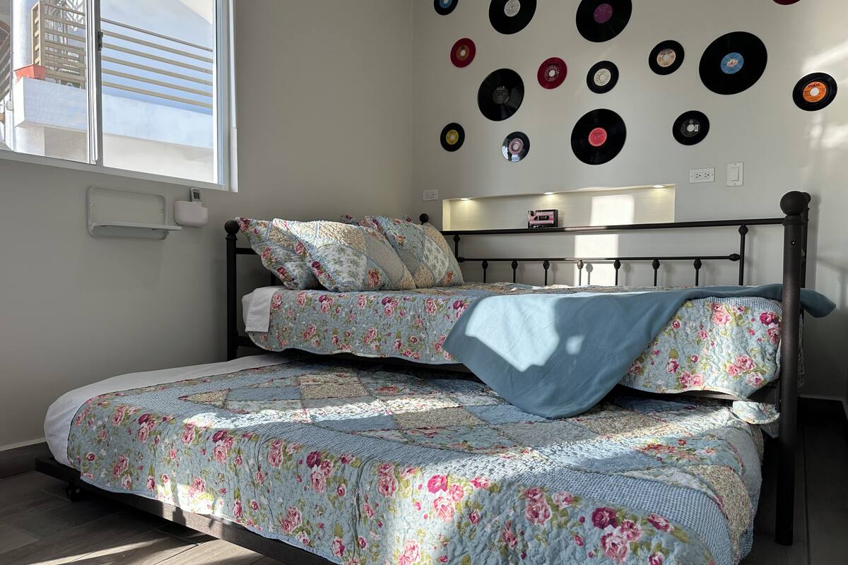 A bed with floral bedding, a wall decorated with vinyl records, and a window with blinds.