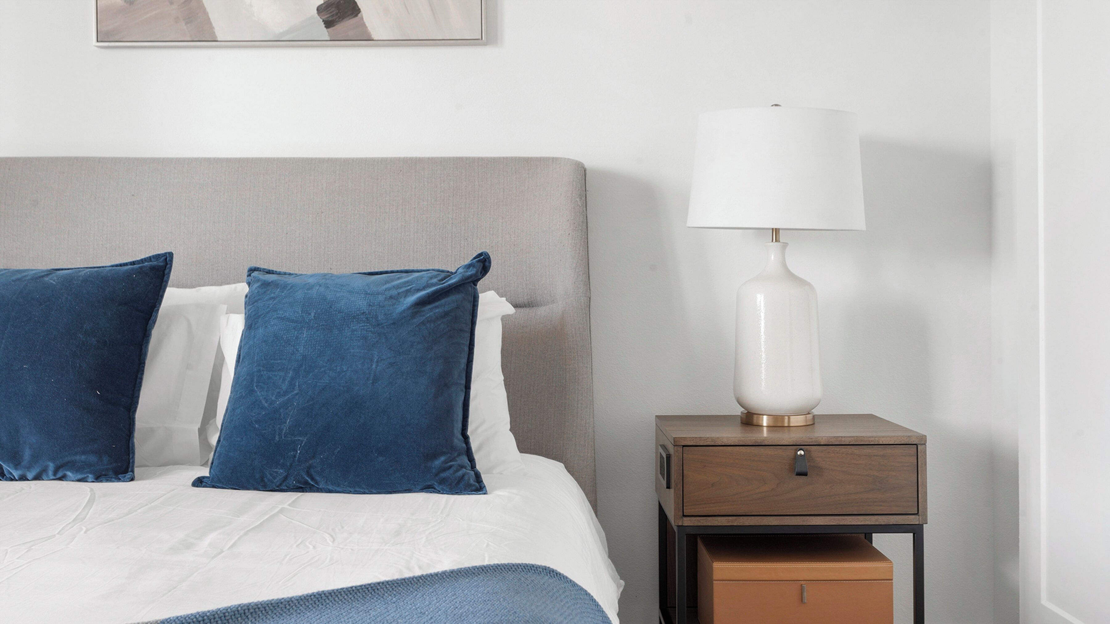 A neatly made bed with blue and white pillows, a white lamp on a wooden nightstand, and a small drawer.