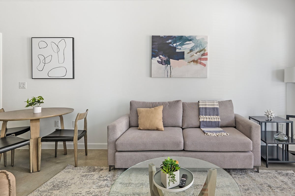 A modern living room with a grey sofa, a round coffee table with a plant, a dining area with a wooden table and chairs, and a painting on the wall.