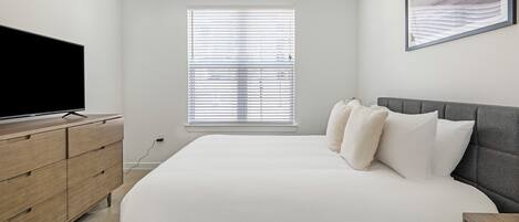 A bedroom with a bed, a TV on a wooden dresser, and a window with blinds.