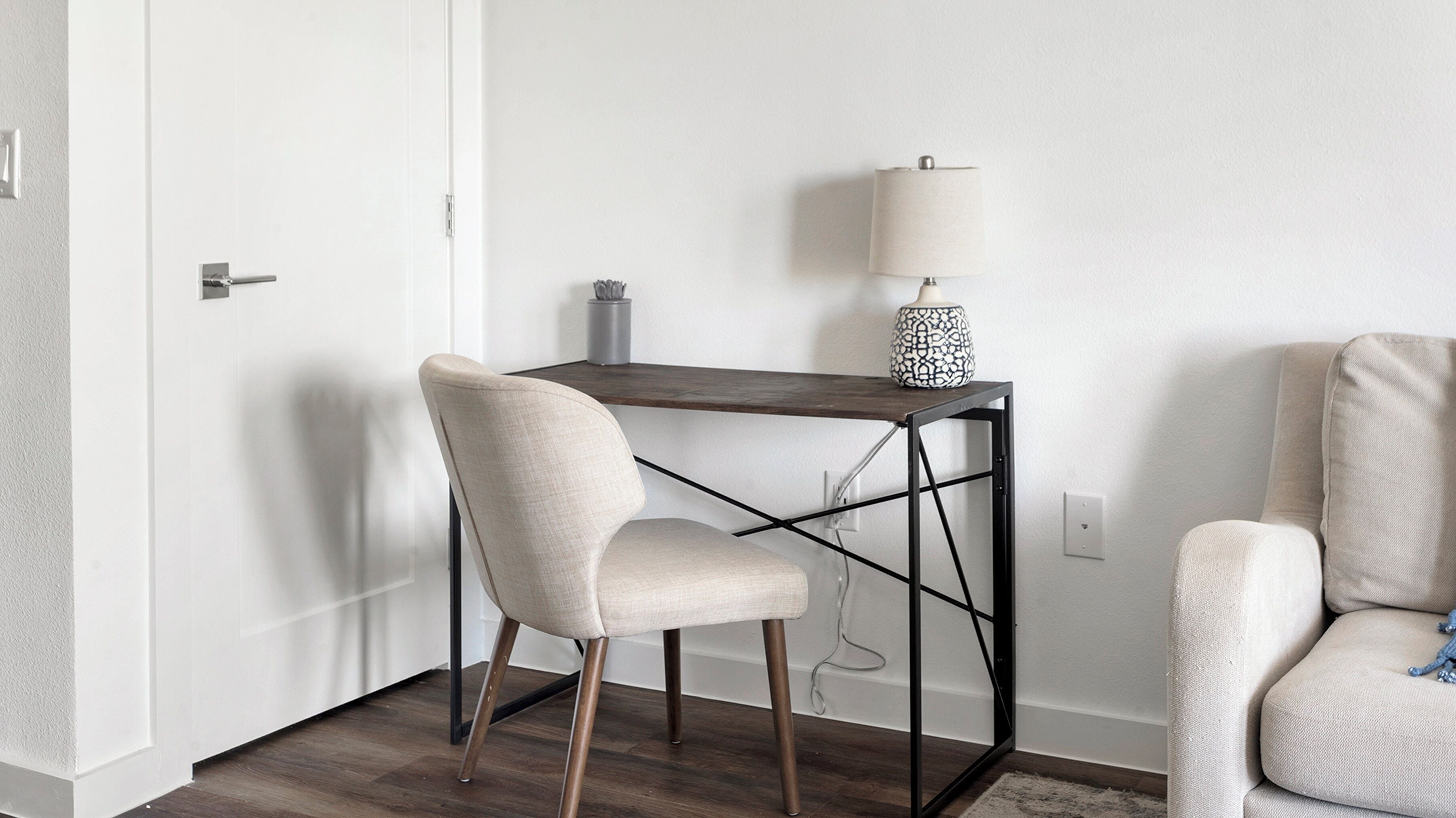 Une petite pièce moderne, équipée d’un bureau et de deux chaises. La pièce est dotée d’une porte blanche, d’une lampe sur le bureau et d’une prise électrique au mur.