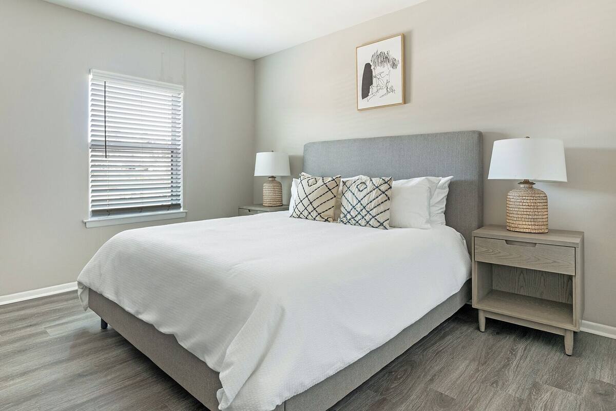 A bedroom with a large bed, two bedside tables with lamps, a window with blinds, and a framed picture on the wall.
