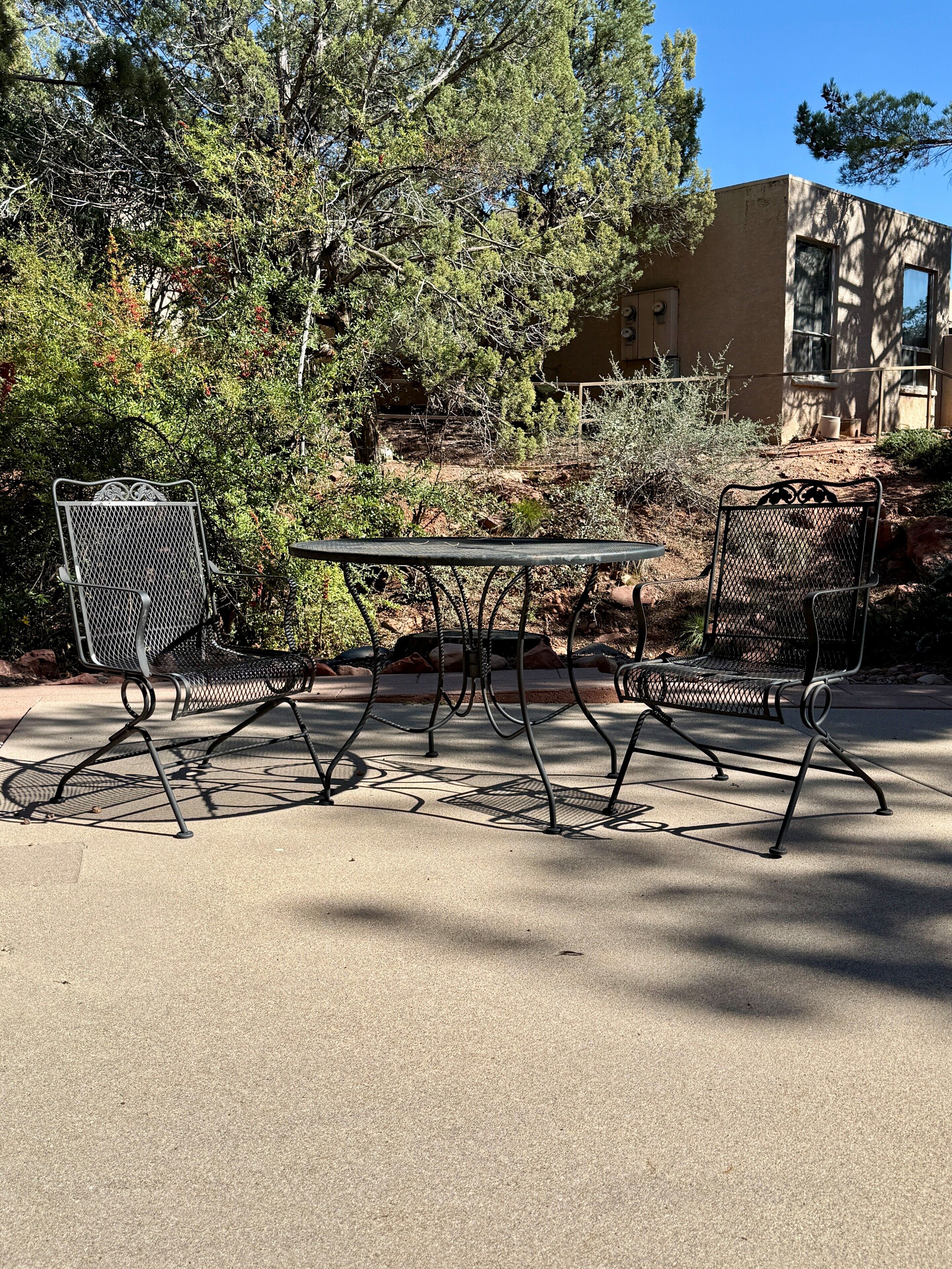 An outdoor seating area with a metal table and chairs, surrounded by trees and a building in the background.