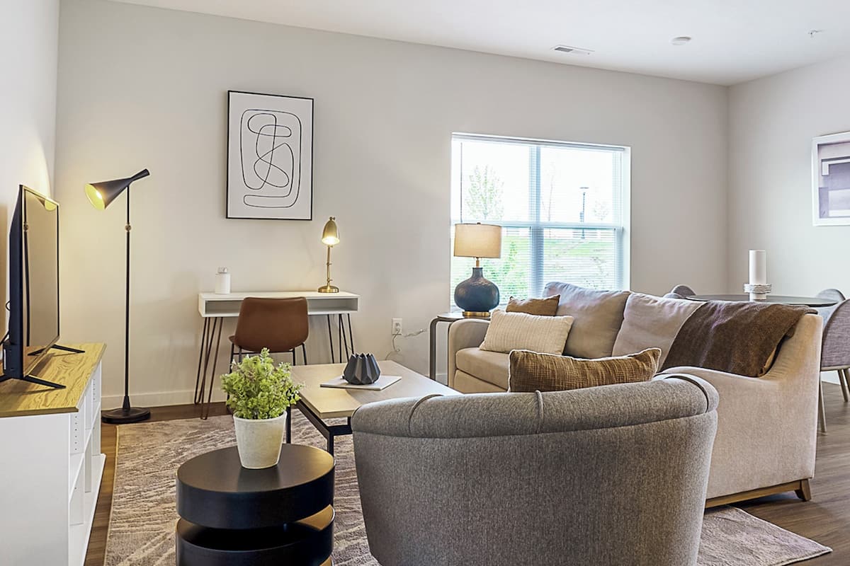 A modern living room with a grey sofa, a coffee table, and a dining area with a table and chairs. There are two wall-mounted lamps and a framed abstract artwork above the sofa.