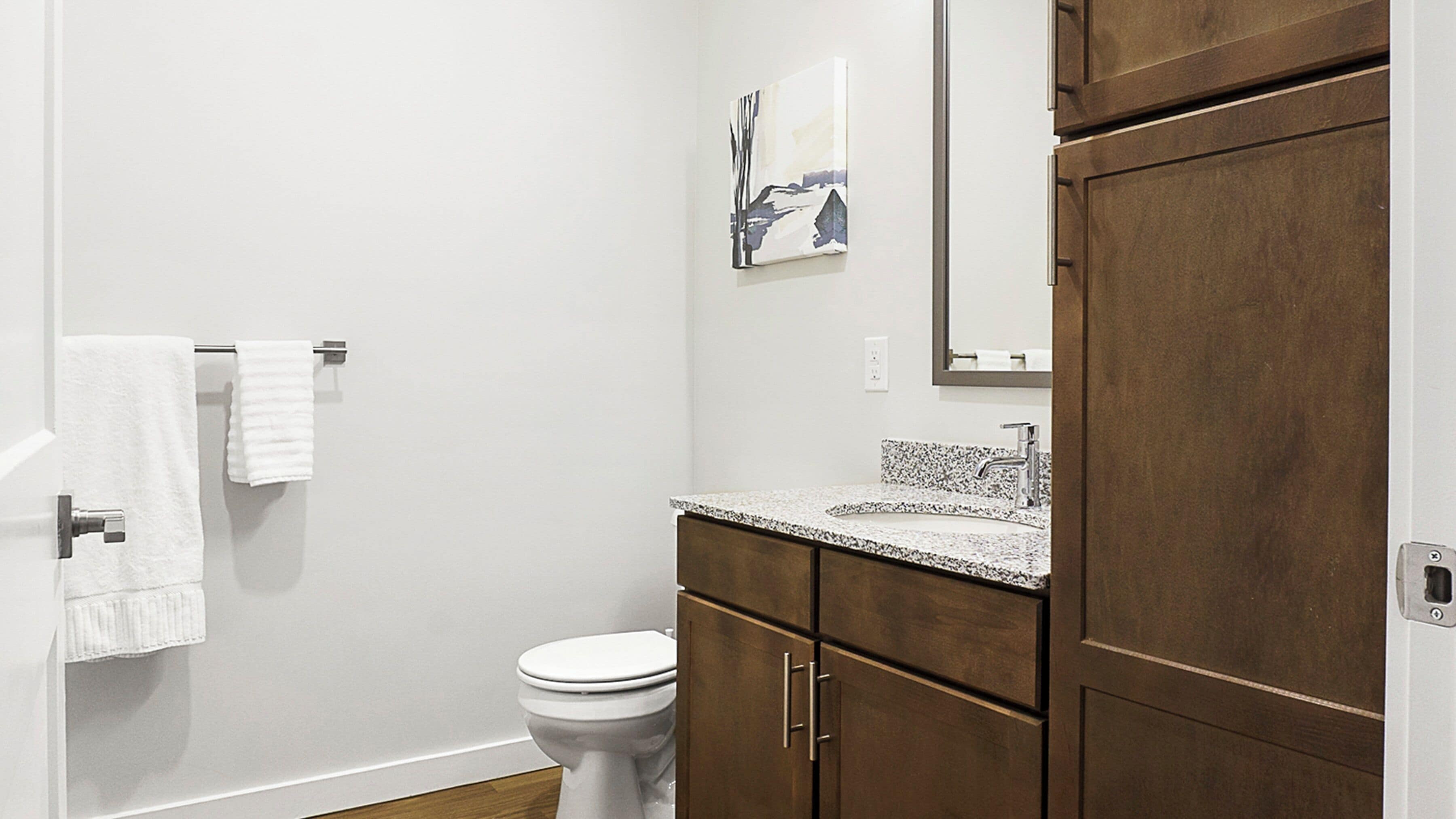 A bathroom with a toilet, a sink with a granite countertop, wooden cabinets, a towel rack with two white towels, and a painting on the wall.