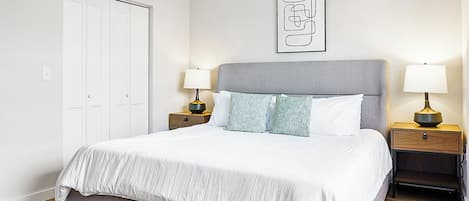 Un dormitorio ordenado con una cama grande, dos mesitas de noche con lámparas, un armario y una obra de arte enmarcada en la pared.