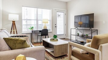 Una sala moderna con sofá, sillón, mesa de centro y televisor sobre un mueble bajo. Hay dos ventanas con persianas, una lámpara y una mesita con plantas.
