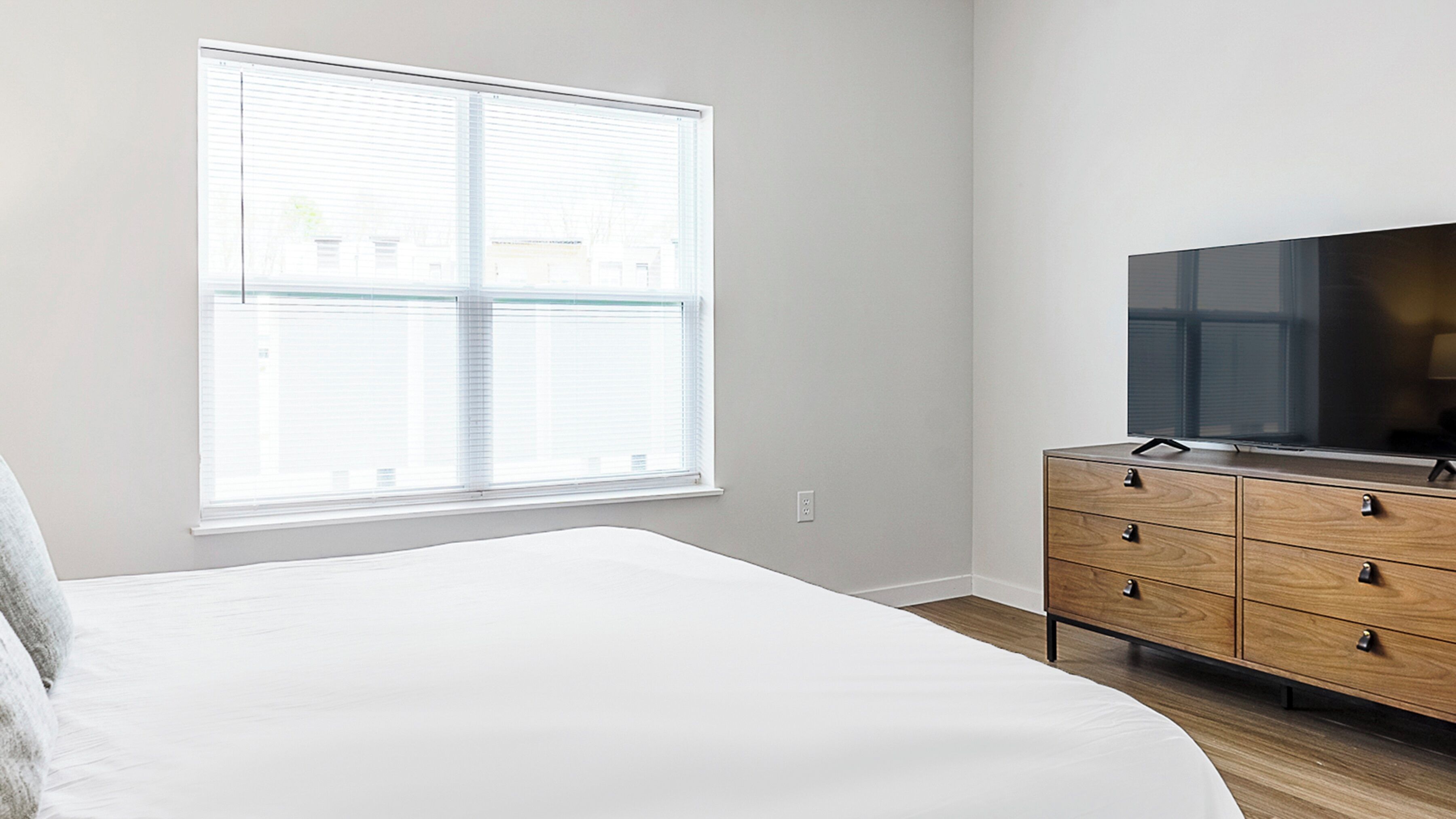 Une chambre à coucher avec un grand lit, une commode en bois, un téléviseur à écran plat et une fenêtre avec des stores.