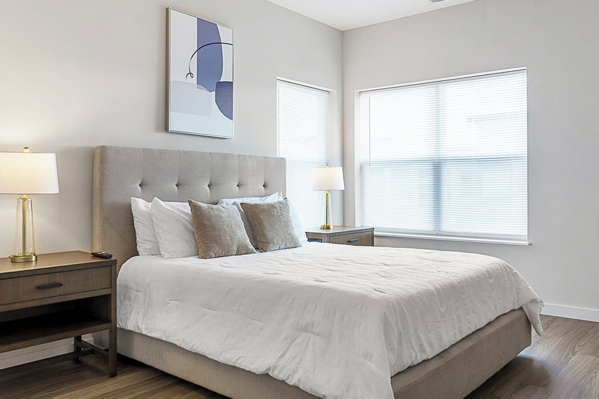 A modern bedroom with a large bed, two bedside tables, a nightstand, and a wall-mounted artwork.
