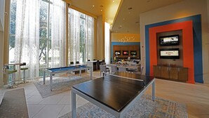 Amplio espacio interior con mesa de billar, mesa de ping pong y taburetes de bar. Grandes ventanales proporcionan luz natural y un televisor montado en la pared.