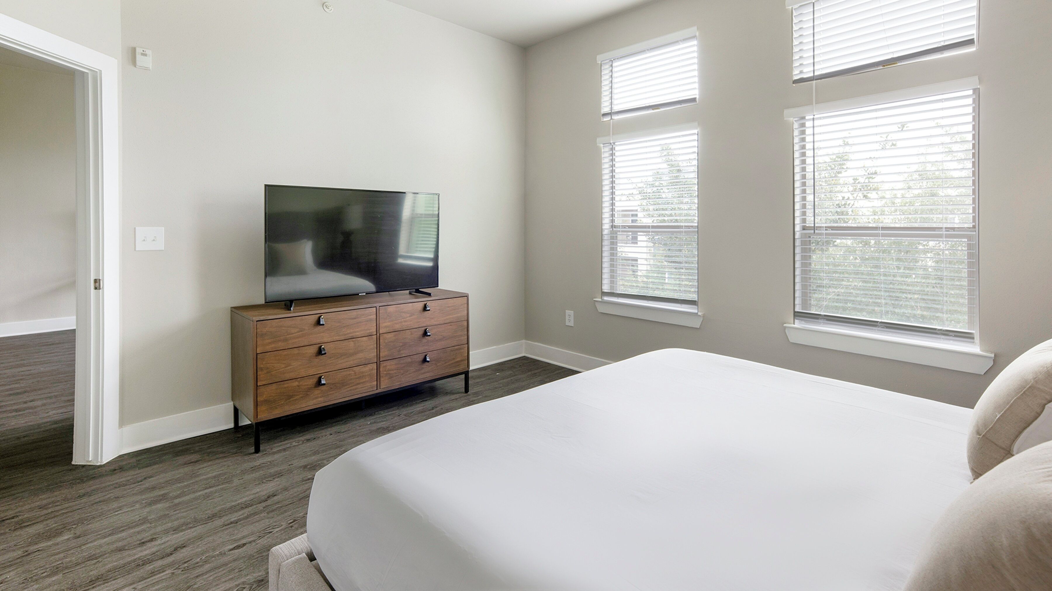 Une chambre avec un grand lit, une commode en bois, un téléviseur à écran plat et trois fenêtres équipées de stores.