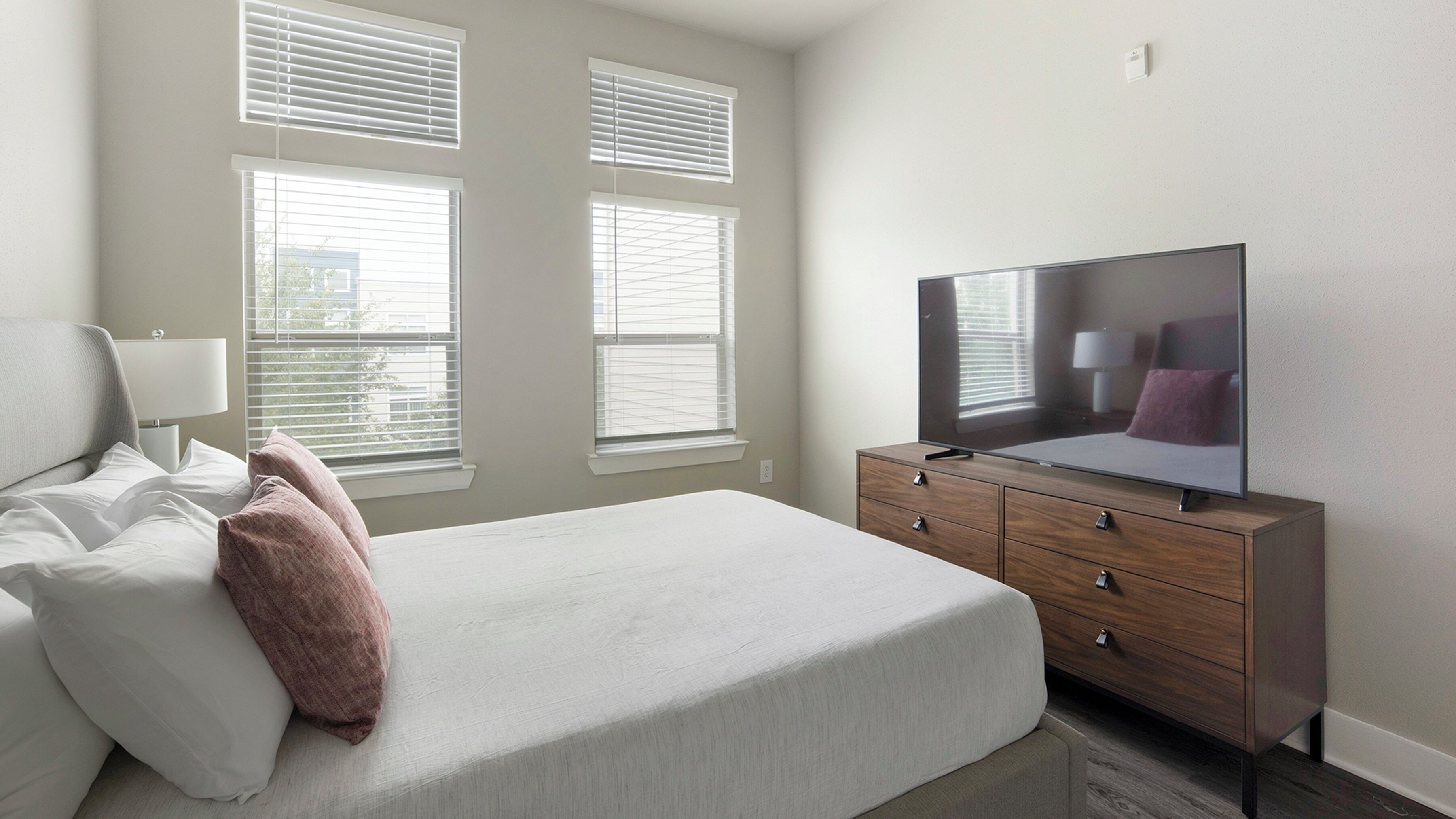 Une chambre à coucher avec un lit, un téléviseur posé sur une commode en bois et des fenêtres équipées de stores.