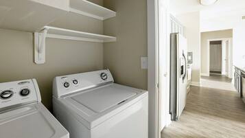 A laundry room with two front-loading washers and dryers, a white shelf above the dryers, and a door leading to a hallway with a kitchen area.