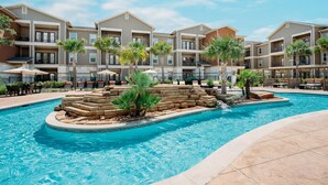 Una piscina con una fuente de piedra y palmeras, rodeada por un edificio de varios pisos con balcones.