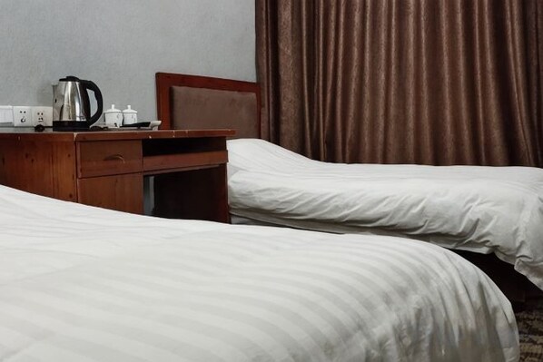 Habitación de hotel con dos camas, una mesita de noche de madera con una tetera y una taza, y una cortina marrón.