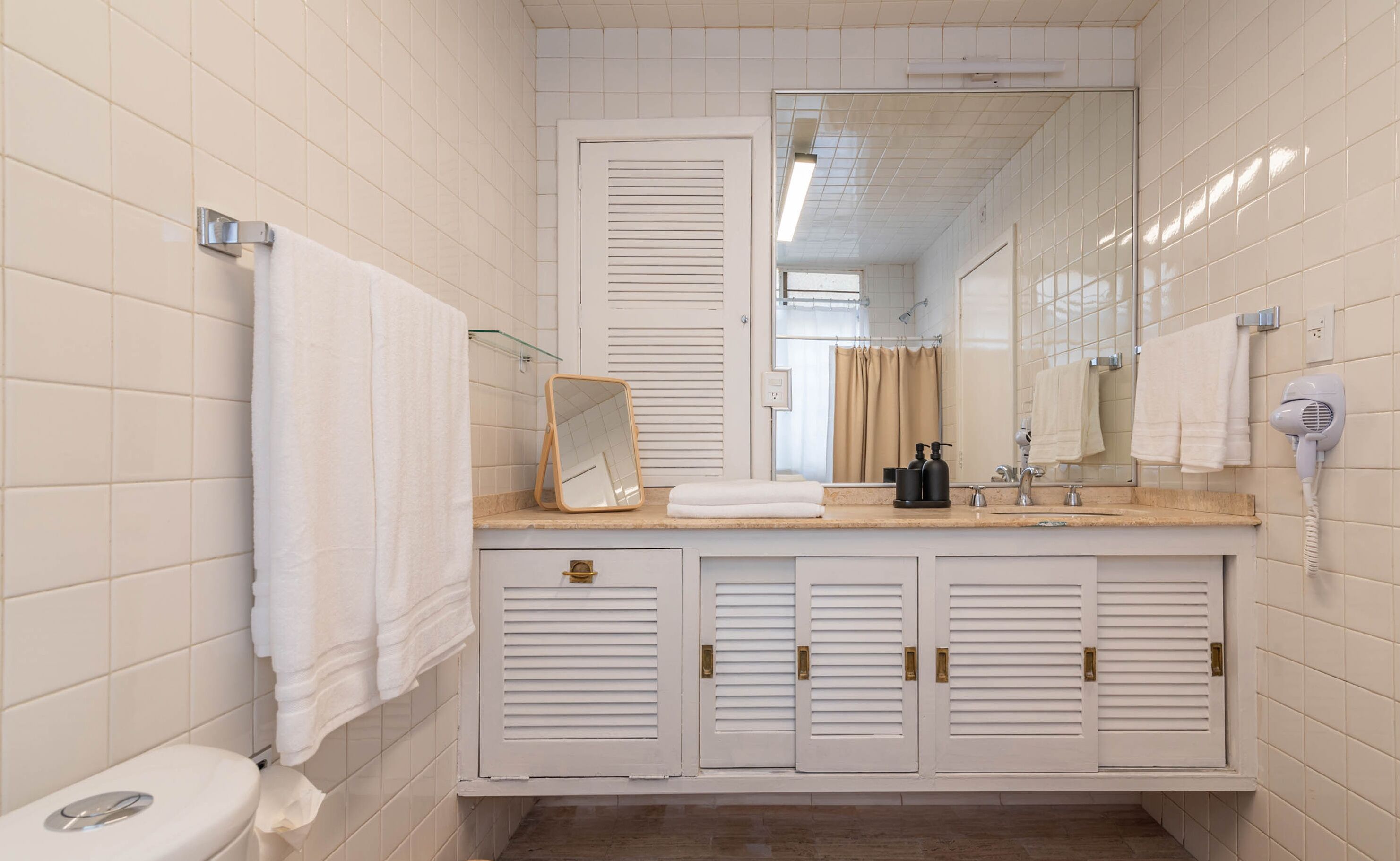 Un baño con paredes de azulejos blancos, un inodoro, un lavamanos con espejo y un secador de pelo.