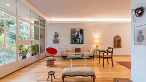 Een ruime woonkamer met een groot raam, een rode fauteuil, een bank, een houten salontafel en een lamp.